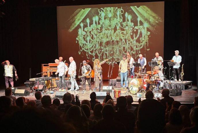 The Band plays on stage as a lively crowd stands in front, cheering, singing, clapping, and enjoying the music together.