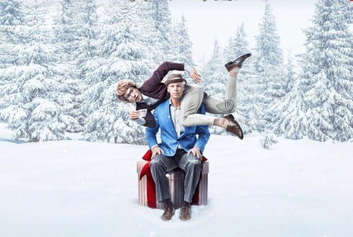 Two people pose playfully in snowy woods, one draped over the other's shoulders while seated on a wrapped gift box.