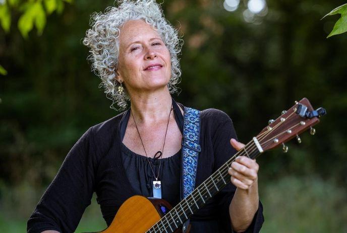 Brenda McMorrow smiling, playing her guitar in a green nature setting.