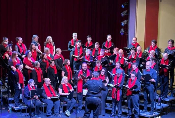 Choir in black attire with red scarves performs on risers as a conductor leads on stage.