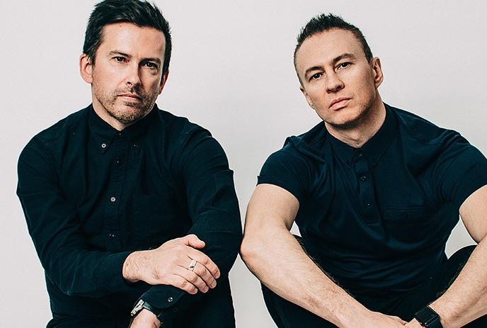 Two men in black shirts sit against a background blending abstract art and earthy textures.