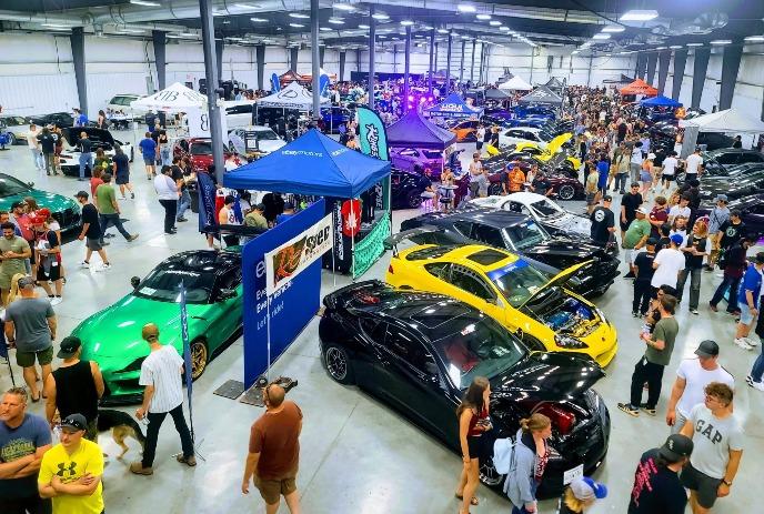 Crowded car show with colorful modified cars and people browsing booths inside a large indoor venue.