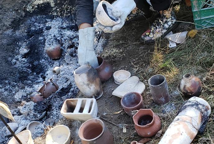 A potter in gloves and floral boots handles pottery near a pit with ashes, surrounded by scattered ceramic pieces