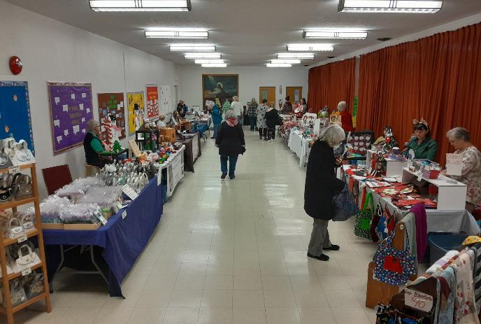 Indoor craft fair in community hall with tables displaying handmade goods, people browsing, and bright lighting.