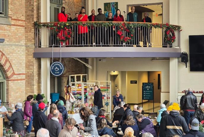 Festive indoor market with crafts, and crowd; balcony singers above, warm holiday atmosphere below.