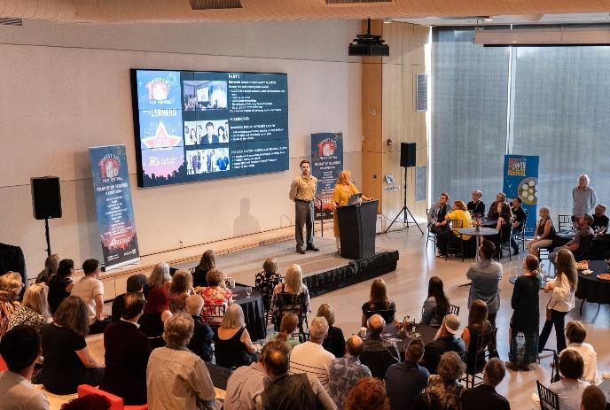 Two people speak at a podium in front of an audience in a conference room with a large screen displaying event details.