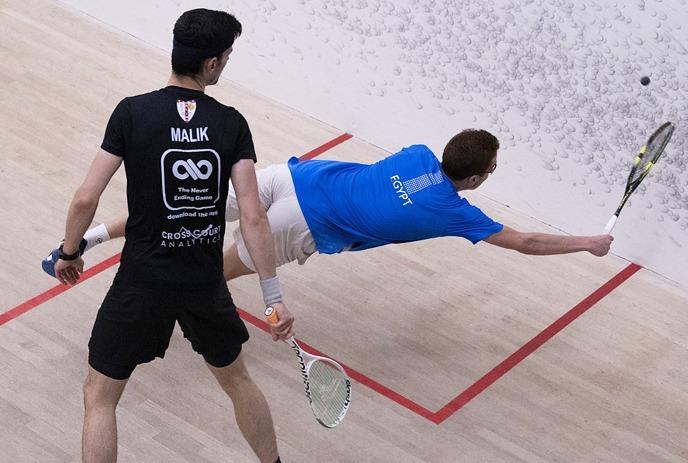 Two squash players in action, one lunging to hit the ball while the other watches.
