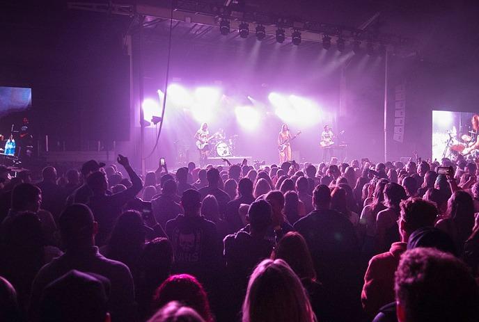 Outdoor concert with band on stage, audience seated under tent, large screen left, music notes graphic bottom left.