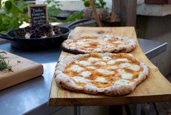 Two rustic pizzas on a wooden board beside a pan of roasted toppings in an outdoor cooking setup.