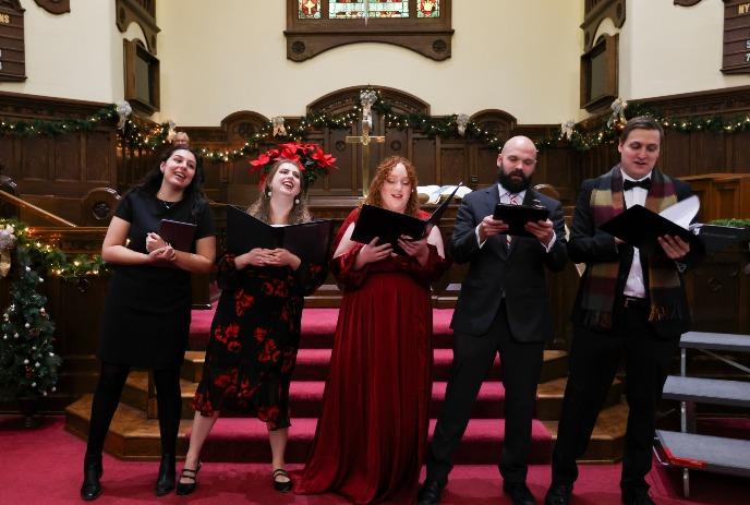 Festive choir sings on a church stage decorated with holiday garlands, standing before a wooden altar.