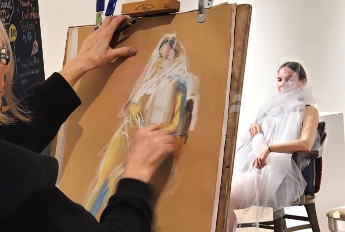 Artist sketching draped model in studio capturing soft fabric folds and seated pose.