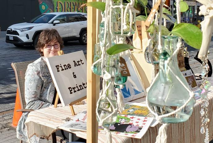 Outdoor art stall with prints and hanging glass bottles in a lively street scene.