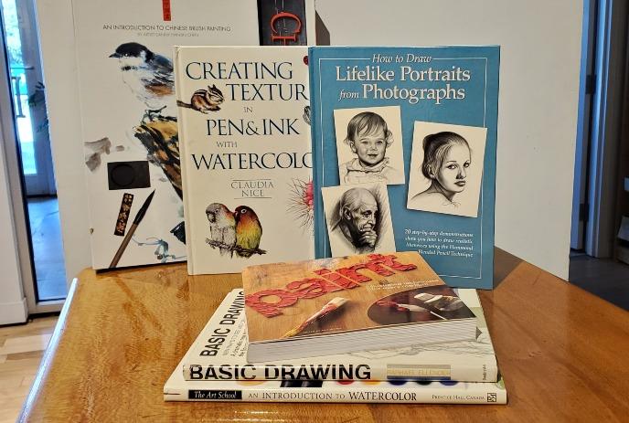 Stacked art instruction books covering drawing painting texture portraits watercolor and brush techniques.