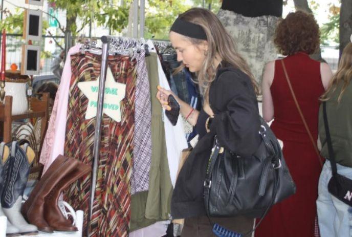 Women browsing vintage clothes at outdoor market rack with patterned items and boots nearby.