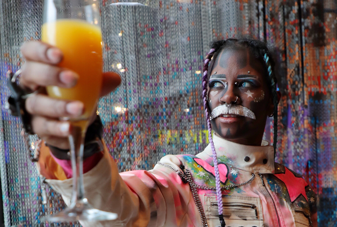 A man wearing colourful clothing, holding up a mimosa in the air, smiling.