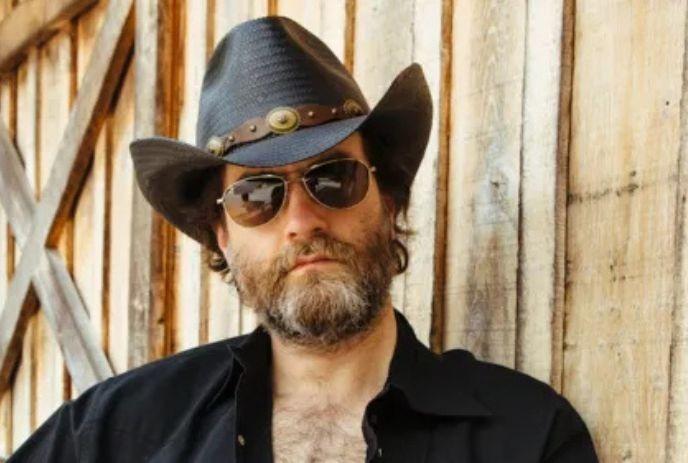 Wheeler Walker Jr. wearing a black cowboy hat, aviator shades and a beard, against a weathered wooden wall.
