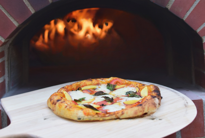 Golden-crusted pizza topped with cheese, tomatoes, and basil is pulled from a glowing brick oven on a wooden peel.