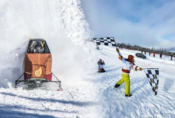 A snowmachine racer powers through flying powder as a jubilant flag-waving woman in bright gear celebrates on the snowy slope.