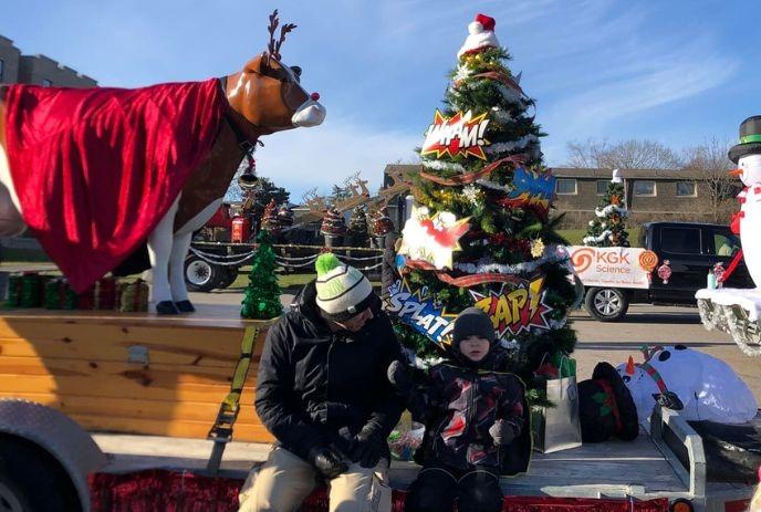 Festive parade float with reindeer, comic-decorated tree, gifts, and two people enjoying holiday fun.
