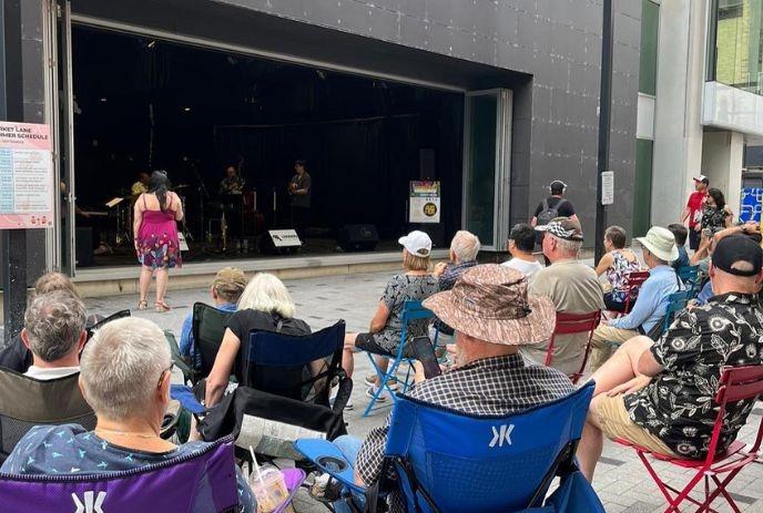 Crowd seated outdoors watching live band perform on small stage between tall urban buildings.