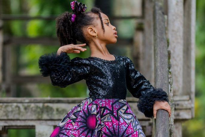 Child in bright patterned skirt and black top stands outdoors near wood with soft green background.