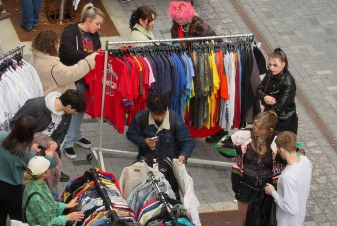 Outdoor market scene with people browsing colorful shirts and jackets on clothing racks.