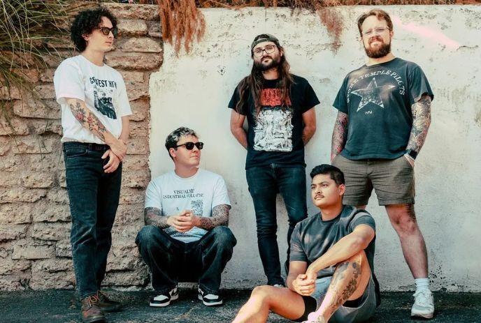Hot Mulligan band poses with neutral, relaxed expressions in t-shirts against a white wall and a textured stone pillar.