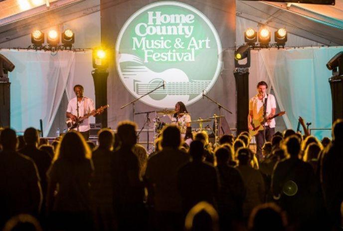 A band performs on a lit stage under a large tent at a music festival with crowd cheering.