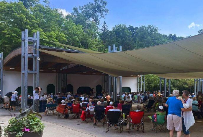 A large crowd sits in an outdoor amphitheater watching a performance on a covered stage