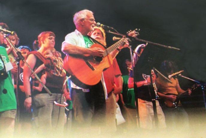 Old photo of Bernie Gilmore playing guitar on a stage with a group of people behind him.