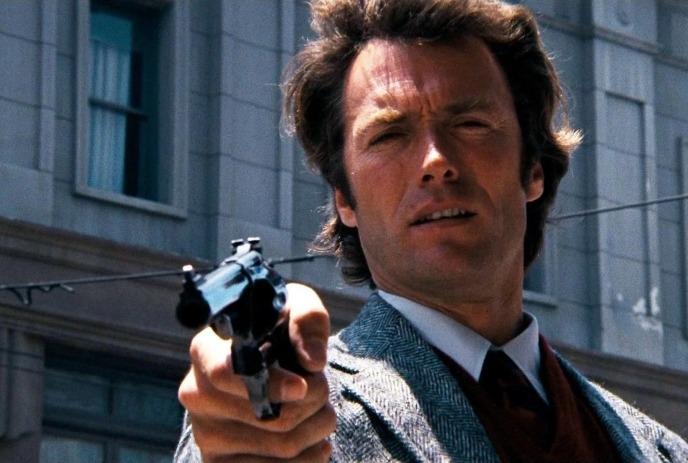 Man in suit points a revolver toward the camera, standing outside a building in a tense scene