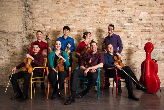 Eight musicians pose with string instruments near a brick wall, featuring a striking red cello case at the right.