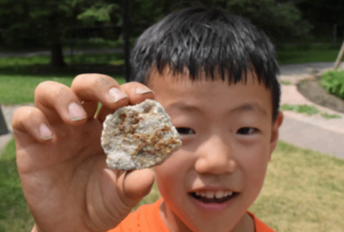 Young child wearing an orange shirt, holding a rock in front of him for everyone to see.