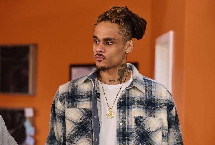 Jo Dolo posing for the camera in front of an orange background, wearing a flannel shirt and chain.
