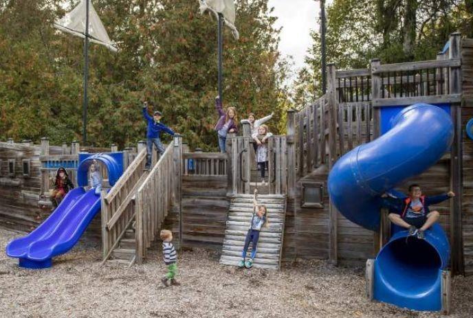 Children playing on a wooden ship themed playground with slides and decks surrounded by trees outdoors.