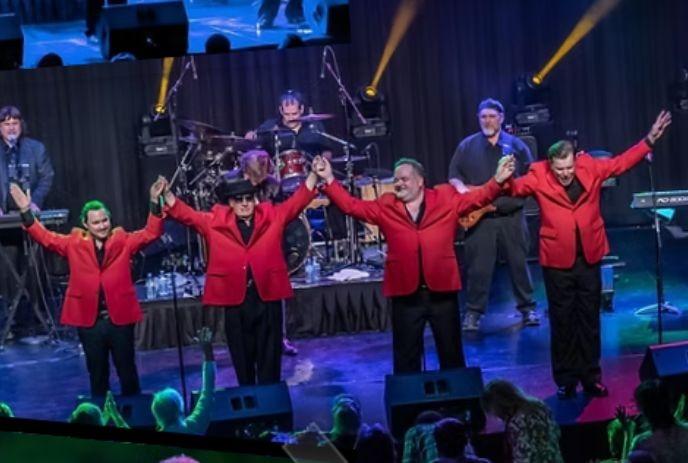 A band group in red jackets holding hands up on a stage, cheered by a crowd.