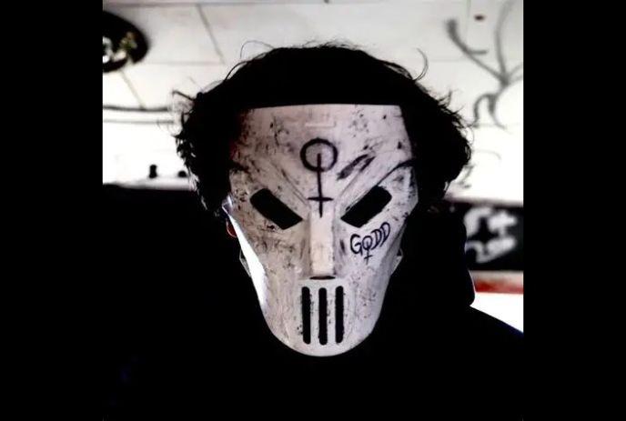 Lil Godd wearing a distressed white mask with black eye slits, hand-drawn symbols, and graffiti-style markings.