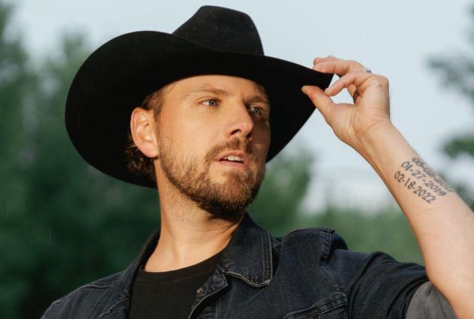 Brett Kissel with a beard and black cowboy hat, tipping his hat with a tattooed arm visible.