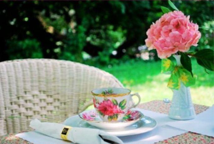 Soft garden tea scene with floral cup white napkin gold ring and pink bloom on table green garden beautys