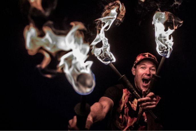 Artist holds three flaming torches in dramatic pose; dark background highlights fire and intense energy.