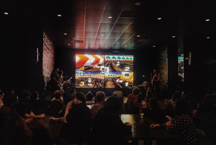 Group of people sitting in front of a large screen with Mario Kart playing.