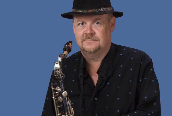 Chris Murphy looking at the camera, wearing a hat and holding a saxophone, in front of a blue background.