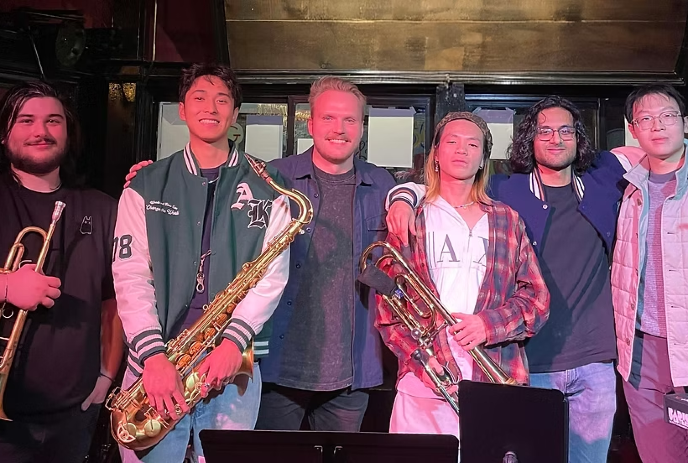 A group of six smiling friends stand close together in a warm, dimly lit room.