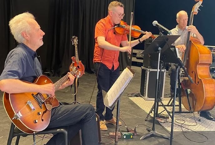 Three musicians perform on stage: a guitarist seated, a standing violinist in an orange shirt, and a double bassist.