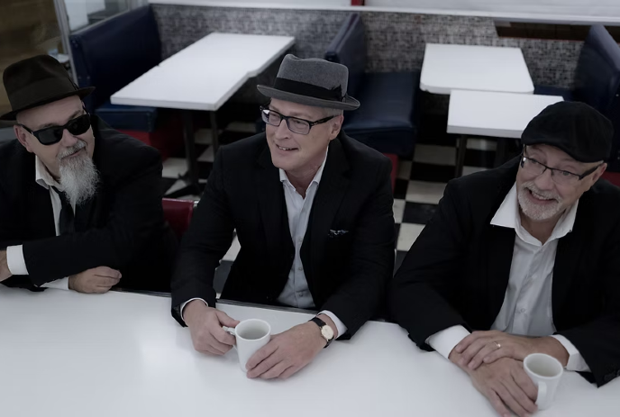 Three men in suits and hats, smiling and holding coffee mugs, sit at a diner counter with black-and-white checkered floors.