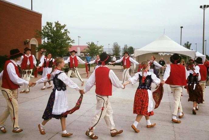 People in folk costumes dancing in a circle outdoors, holding hands, with a tent and buildings behind them.
