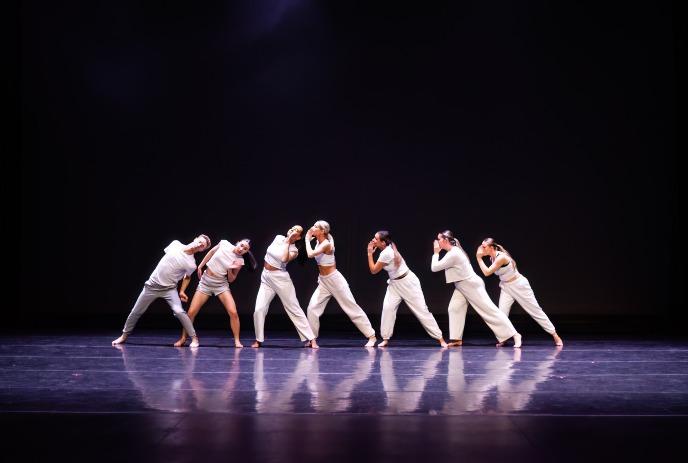 Seven dancers in white outfits perform a synchronized routine onstage under dramatic spotlight illumination.