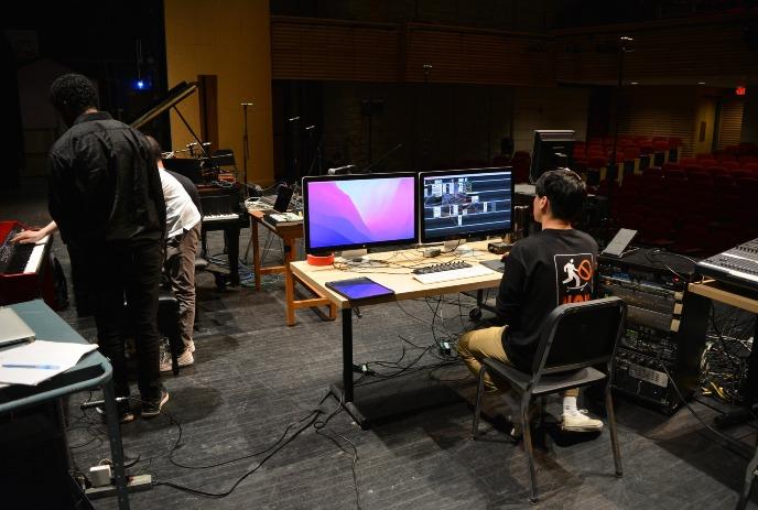 Technicians and musicians prep stage with piano, monitors and theater seats in the background.
