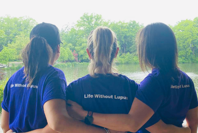 Three women wearing matching 'Life Without Lupus' shirts with their hands around each other, looking at a body of water.