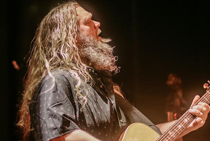 Matt Andersen with long curly hair and beard playing an acoustic guitar under warm stage lighting.